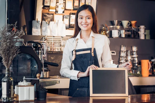 Asian female barista wear jean apron and holding blank blackboar
