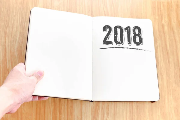 Hand holding open notebook with 2018 new year lay it on wooden t — Stock Photo, Image