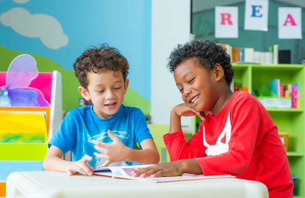 Dos niños se sientan en la mesa y leen libro de cuentos en preescolar lib —  Fotos de Stock