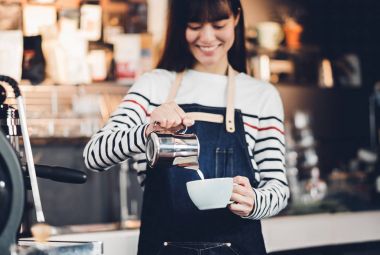 Asya kadın barista sayaç barda sıcak kahve fincan içine süt dökün 