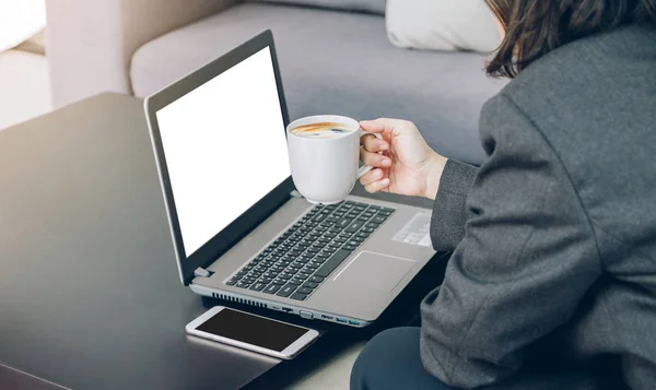 Donna d'affari in possesso di una tazza di caffè che lavora su laptop e cellulare a — Foto Stock