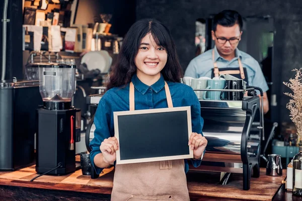 Asian female barista wear jean apron holding blank blackboard co