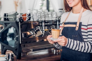Asya kadın barista servis sıcak kahve fincanı tutan jean önlük giymek