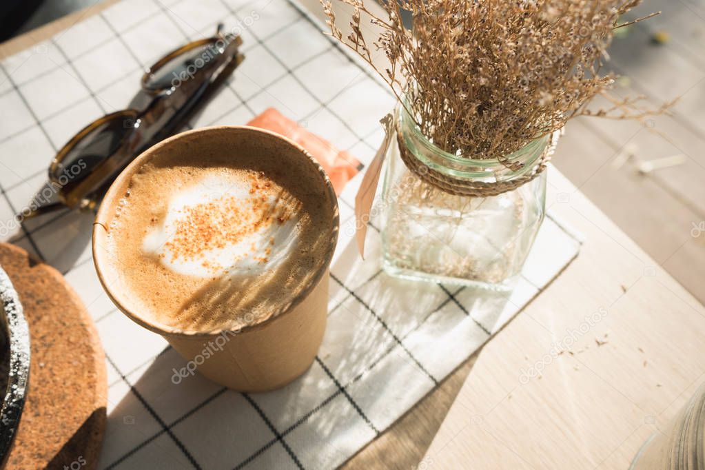 cappuccino in take away coffee cup on table cloth with dry flowe