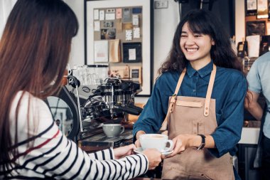 Asyalı kadın barista kahve fincanı için hizmet tutan jean önlük giymek