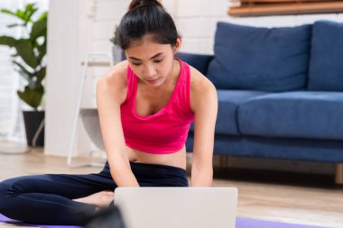 Asian woman in sportswear sitting on matt in living room and tra