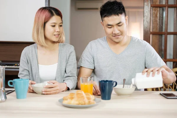 happy asian couple in pajamas sitting at table in kitchen at home in morning and having cereal breakfast together