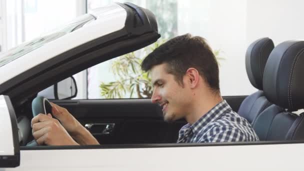Glücklicher junger gutaussehender Mann sitzt in einem neuen Cabrio im Autohaus — Stockvideo