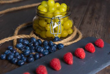 Bodegon meyvesi, levhada taze çilek, yaban mersini ve yıllanmış üzümlerle dolu cam kavanoz.