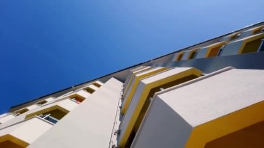 Alttan binanın balkonlarında açık mavi gökyüzüne karşı sarı çizgiler var.