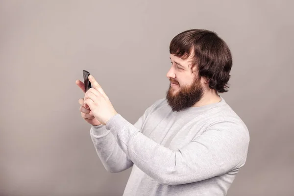 Vista lateral del joven hombre guapo con camisa gris claro tomando fotos con teléfono móvil aislado sobre fondo oscuro del estudio . Imágenes de stock libres de derechos