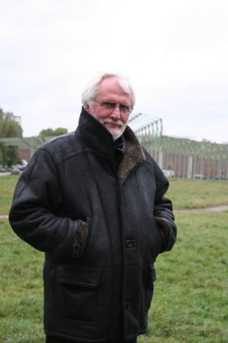 Beyaz sakallı, gözlüklü ve uzun siyah deri ceketli yaşlı adamın portresi parkta duruyor.