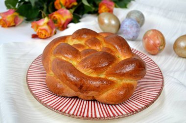 Taze pişmiş challah / maya rulo / brioche / tatlı hamur örülmüş Paskalya yumurtaları beyaz masa örtüsü ve arka planda güller 