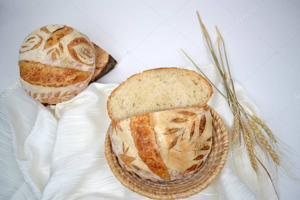 Two loafs of home baked  crusty sourdough bread; one cut in half in bread basket, another loaf on wood; wheat ears and white table cloth in background 