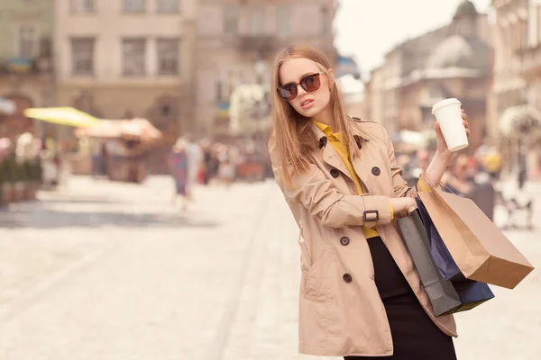 Shopping therapy. Young fashionable woman taking a coffee break after shopping, walking with a coffee-to-go in her hands against urban city background. — Stock Photo, Image