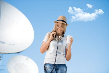 Mutlu genç kadın vintage müzik kulaklık boynuna takmış, paket servisi olan restoran kahve içme ve Kablosuz sinyaller uydulardan aldığı parabolik çanak çerçevede poz şapkalı.