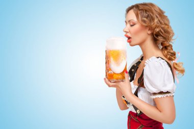 Kırmızı jumper şort ile geleneksel bir dirndl şeklinde jartiyer giymiş genç susuz seksi İsviçreli kadın kupa mavi zemin üzerine dışarı akan köpük ile bira içmek.