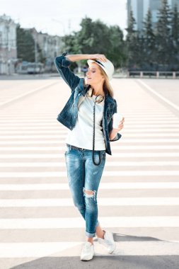 Zebra geçiş ile yolda duran, kahve içme ve Kentsel şehir arka karşı dans tek kullanımlık kahve fincanı, şapka ve kot pantolon ceketle güzel genç kadın.