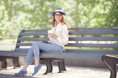 Tablet pc ve bir yaz parkta bankta oturmuş bir tek kullanımlık kahve fincanı ile mutlu bir genç öğrenci.