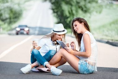 Müzik kulaklık, tablet kullanma ve yol arka plan karşı çevrimiçi müzik satın alma giyen bir paket servisi olan restoran kahve fincandan kahve içme yolda oturup mutlu genç kadın.