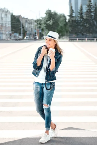 使い捨てのコーヒー カップ 横断歩道で道路の上に立って コーヒーを飲みながら 都市の背景にダンスと帽子とジーンズのジャケットの美しい若い女性 — ストック写真