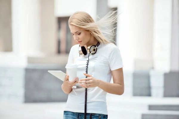 幸せな若い女性彼女の首にビンテージ音楽ヘッドフォンで取る離れてコーヒー カップを置くと都市背景にタブレット でインターネットをサーフィン — ストック写真