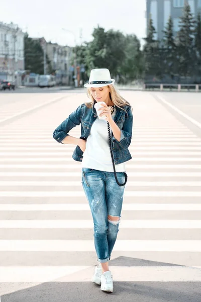 使い捨てのコーヒー カップ 横断歩道で道路の上に立って コーヒーを飲みながら 都市の背景にダンスと帽子とジーンズのジャケットの美しい若い女性 — ストック写真