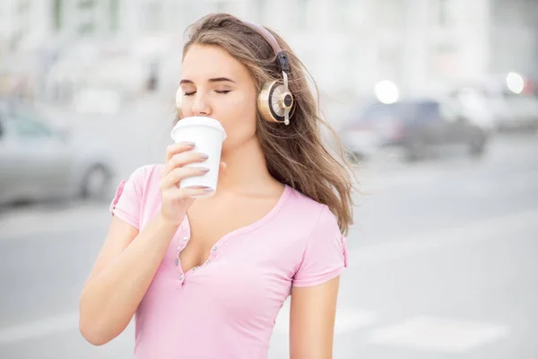 美丽的年轻女子 与音乐耳机 捧一拿掉咖啡杯和听音乐与她的眼睛闭市交通背景 — 图库照片