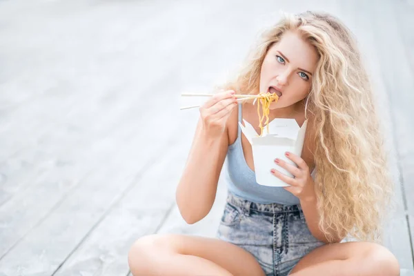 長い巻き毛の木の床の上に座って ランチ ボックスを持って 中国のテイクアウトから麺を箸で食べて幸せな若い女 — ストック写真