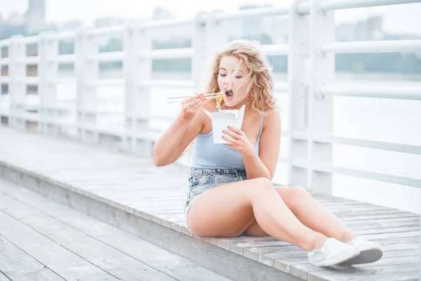 長い巻き毛の木の床の上に座って ランチ ボックスを持って 中国のテイクアウトから麺を箸で食べて幸せな若い女 — ストック写真