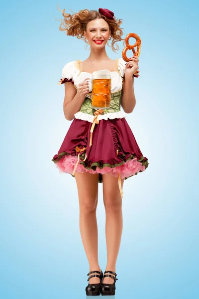 年轻性感的慕尼黑啤酒节服务员 穿着传统的巴伐利亚服饰裙和服务在蓝色背景上的一个小酒馆椒盐脆饼和啤酒的杯子 — 图库照片
