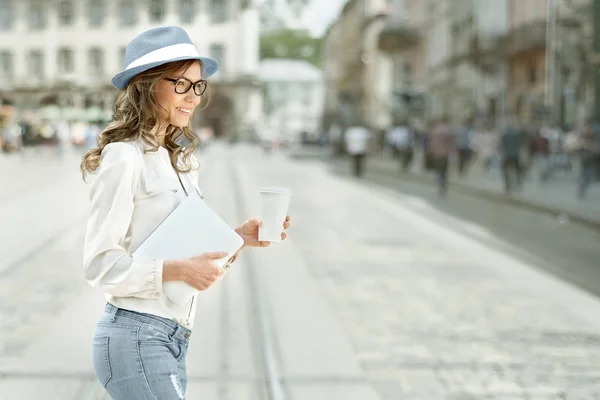 使い捨てのコーヒー カップでコーヒーを飲みながら 彼女の手でタブレットを保持して 背景に都市通りを横断の美しい若い女性 — ストック写真