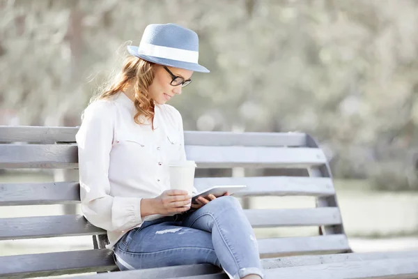 幸せな若い学生タブレットと使い捨てのコーヒーカップベンチに座って夏の公園で読書 — ストック写真