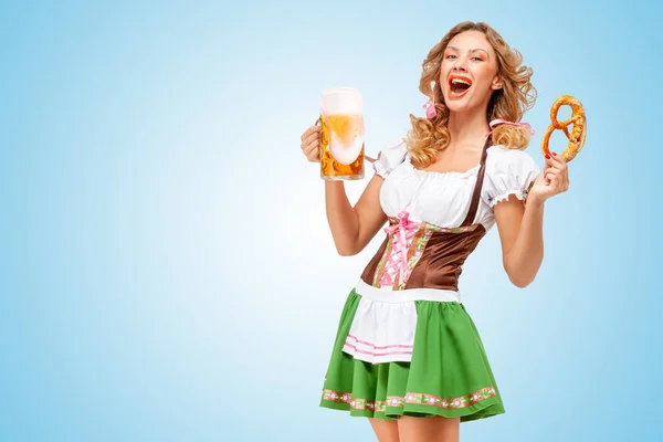 年轻性感的慕尼黑啤酒节女服务员穿着巴伐利亚传统服饰裙提供一个椒盐卷饼和啤酒的杯子 在蓝色背景上 免版税图库照片