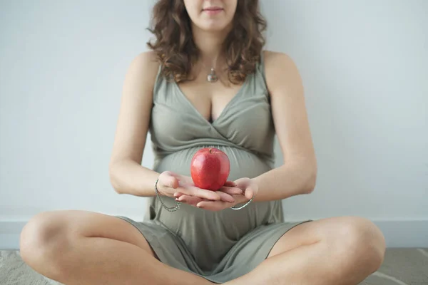 Крупный план обезличенной беременной женщины с большим животом, сидящей на полу и держащей яблоко перед собой — стоковое фото