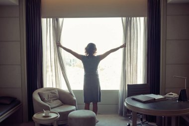 Otel odasının perdelerini açan bir kadının silüeti.