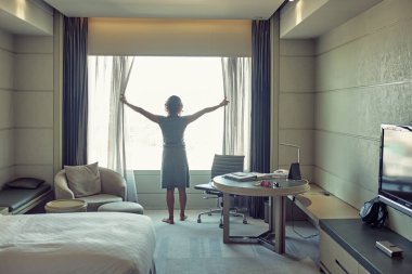 Kadın perdeleri açıyor ve otel odasının penceresinden manzarayı seyrediyor.