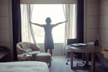 Perdeleri açan ve otel odasının manzarasına hayran bir kadının silueti.