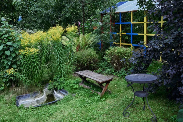 Часть сада с деревянной теплицей ручной работы, скамейкой, столом и обилием зеленых насаждений различных типов. Ландшафтный дизайн . — стоковое фото