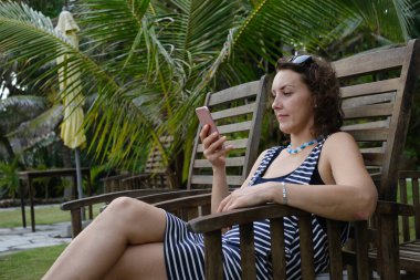 Kahverengi saçlı bir kadın otelin dışındaki tahta sandalyede akıllı telefon kullanıyor..