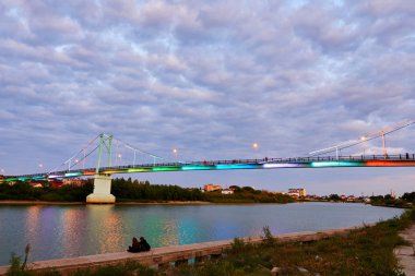 Gün batımı zamanı. Nehrin kıyısında köprü ve yansıtan ışıklarla birlikte.