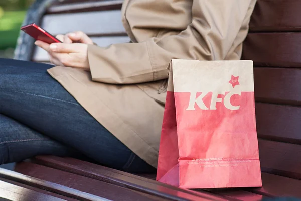 Минск, Беларусь, 21 июня 2017 года: бумажный пакет KFC на скамейке рядом с женщиной с мобильным телефоном — стоковое фото