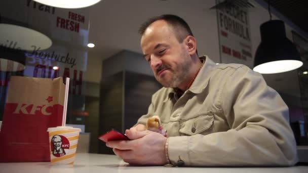 Minsk, Biélorussie, le 5 juillet 2017 : L'homme mange le déjeuner avec beaucoup d'appétit dans un restaurant KFC et en utilisant un smartphone. Vie moderne, dîner avec un smartphone . — Video