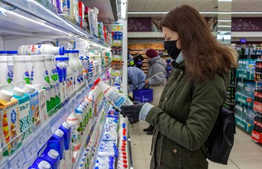 MINSK, BELARUS - 27 Nisan 2020: Koronavirüs salgını sırasında markette koruyucu maskeli bir adam süt alışverişi yapıyor. Malzemeleri, beyanı ya da ürün üzerindeki son kullanma tarihini okuyan genç bir kadın.