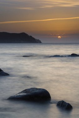 Denizin ortasında, ön planda ve ufukta kayalar ve bulutların ortasında güneşi hayal edin.