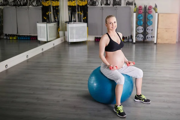 Lächelnde Schwangere sitzt auf Gymnastikball und stemmt Gewichte — Stockfoto