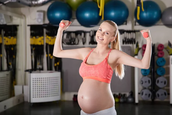 Smiling pregnant woman raising hands with weights at gym