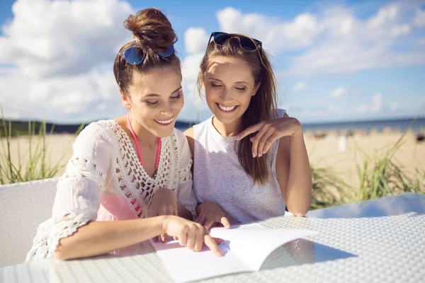 Duas meninas felizes sentadas na mesa de café no menu de leitura da praia — Fotografia de Stock