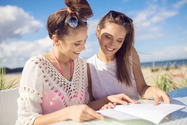 Zwei hübsche Mädchen sitzen am Cafétisch am Strand und lesen Speisekarte — Stockfoto