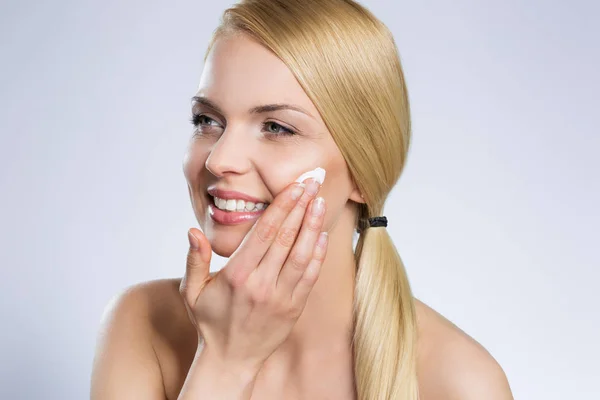 Joven hermosa mujer aplicando crema en la cara mirando hacia otro lado —  Fotos de Stock